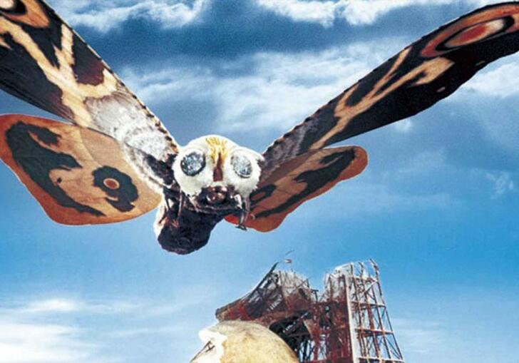 A giant moth creature flies over an amusement park ride that has been smashed.
