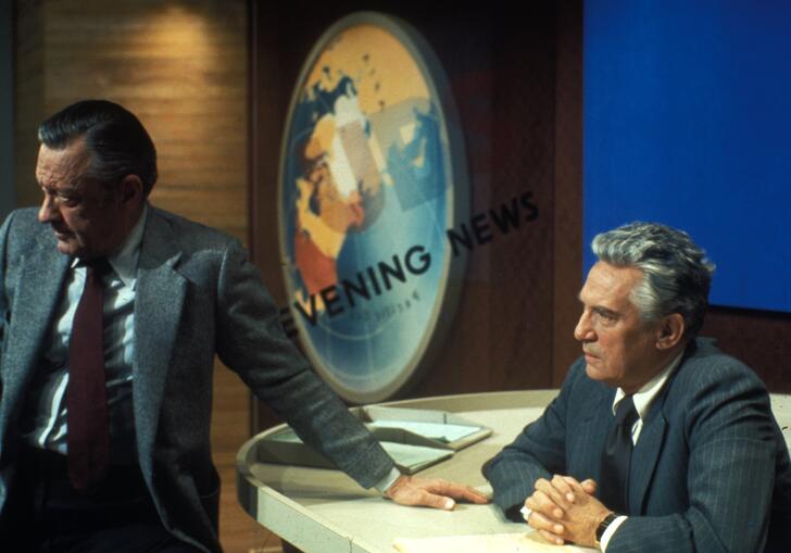 A man sits at a 70s American news broadcast desk. 