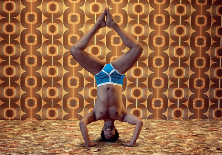A person does a headstand in their pants against an orange and brown patterned wall. 
