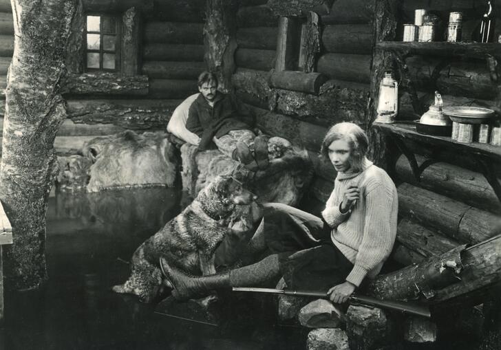A black and white image of a man and a woman sitting by a log cabin with a large dog.