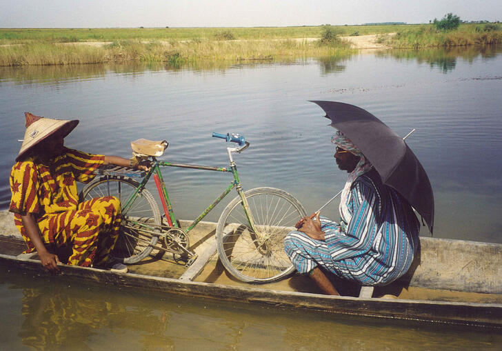 Two old men sit in a long boat on a river with a bicycle and a black umbrella.