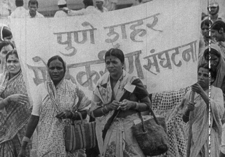 Women stand infront of a protest banner marching down the street.