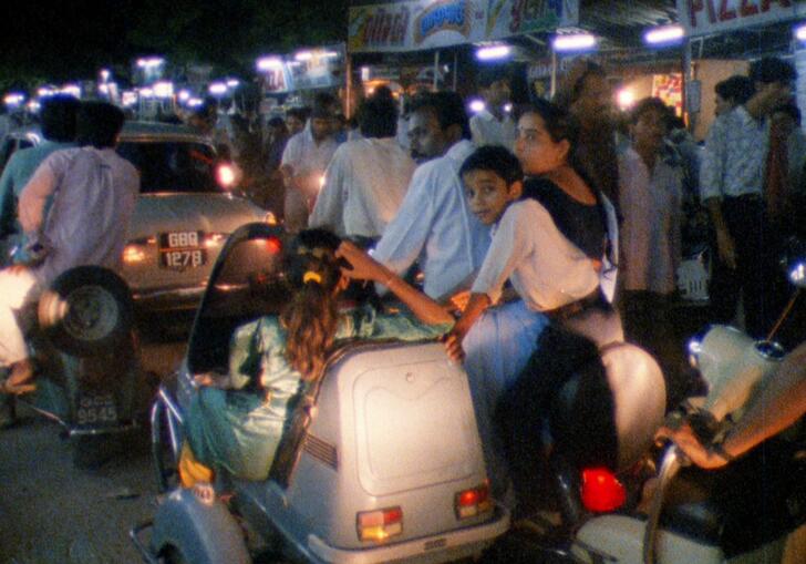 A family sit around a car on an urban street at night.