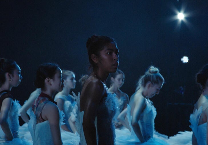 A group of ballet dancers stand in the dark on stage, dressed in feathered white dresses.