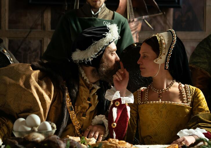 A man and a woman in tudor dress gaze into each other's eyes at a banquet, as the woman touches the man's nose.