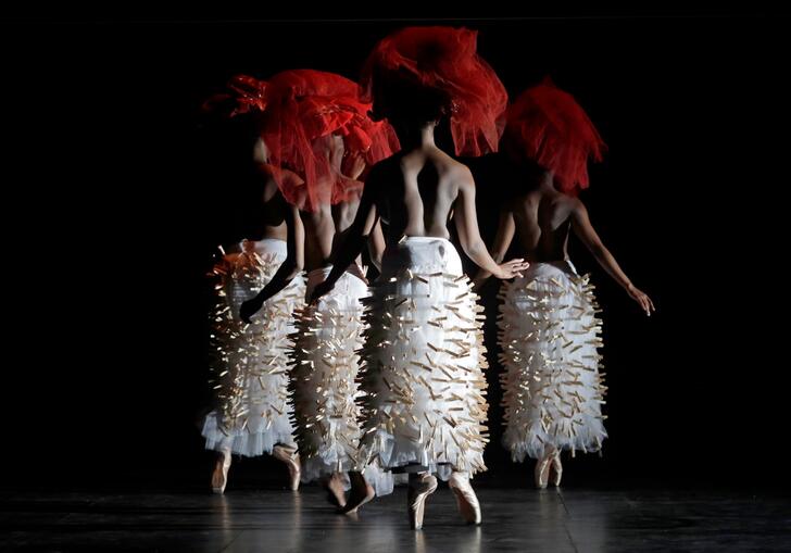 A group of dancers stand on point in a group. They wear red headdresses and white skirts that are decorated with pegs.