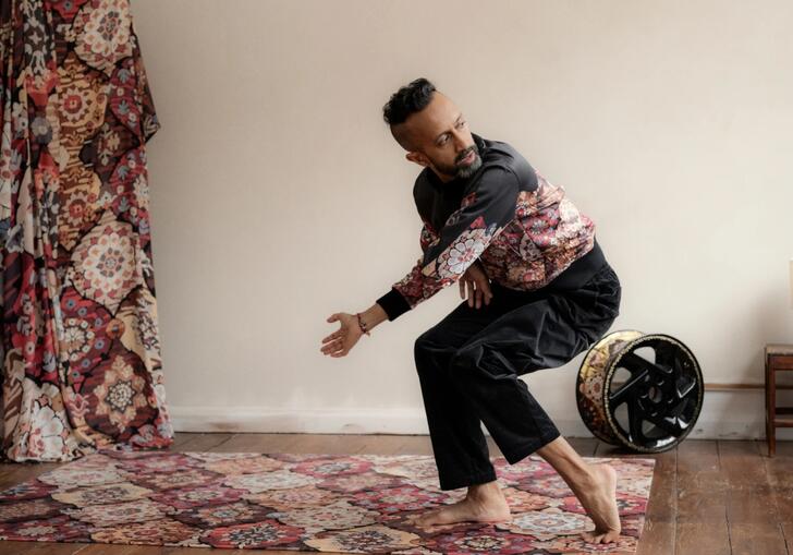 Hetain Patel holds an angular pose while performing on a red fabric in a room with a wooden floor.