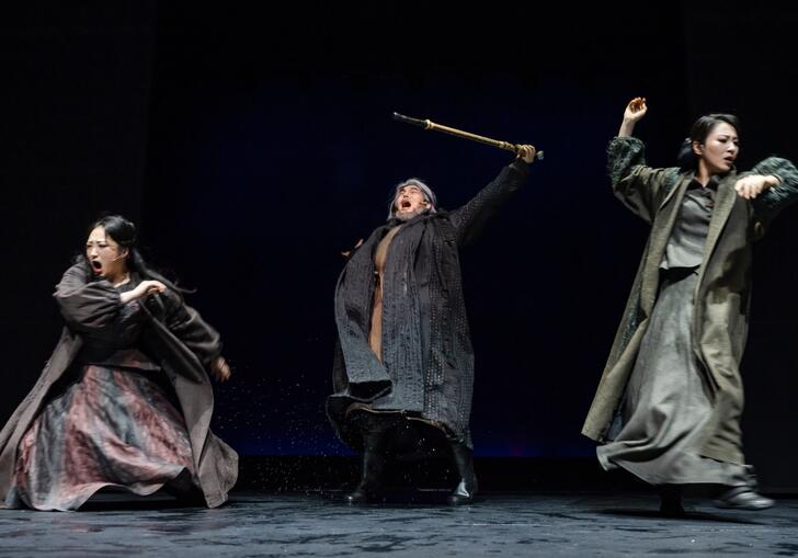 Three performers rage on stage in a production of Lear. They wear muted coloured clothing against a black background.