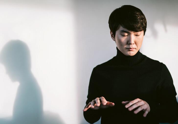 Pianist Seong-Jin Cho in front of a white wall, with eyes closed lifting his hands as if playing the piano