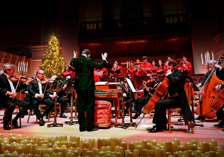 conductor conducting a choir and orchestra behind a row of candles