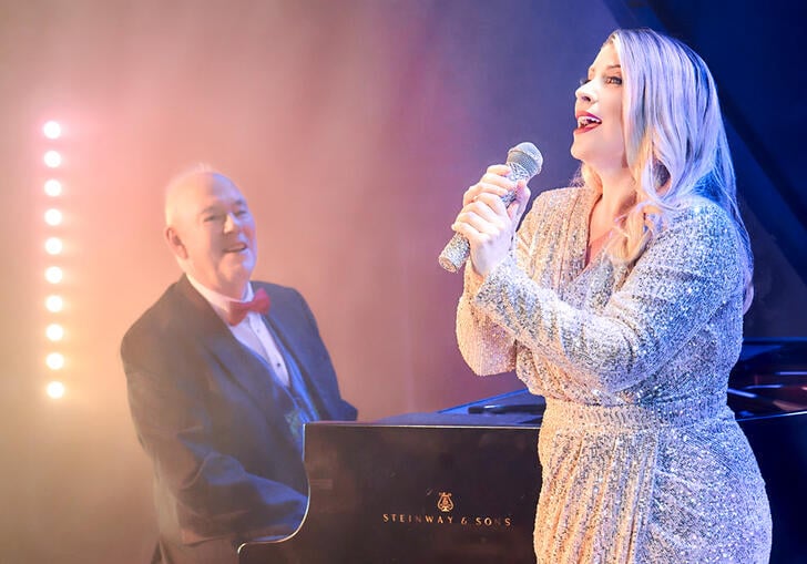 lady in a silver dress singing into a microphone stood in front of a man playing the piano