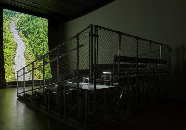 In a dark gallery, metal bleacher-style seating is positioned in front of a large, vertical, curved screen showing a river coursing through a lush, forested valley.