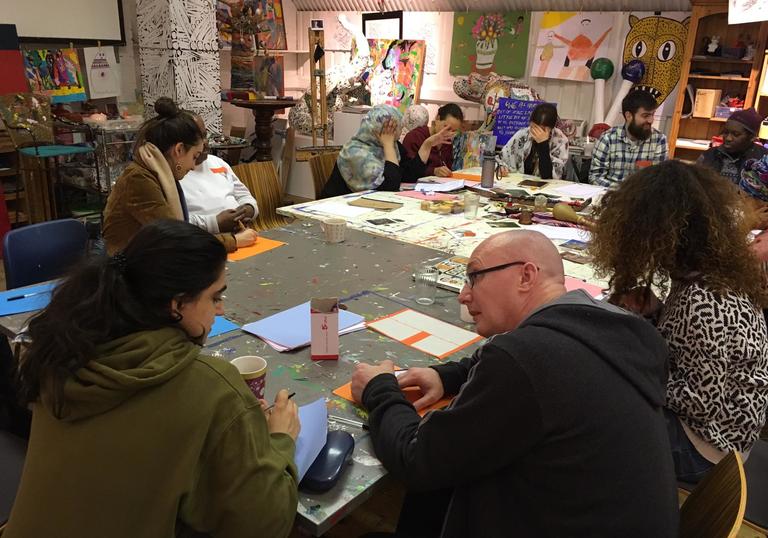 a group sit round an artisit studio table in discussion with one another. It is the Barbican Young Poets and the memebers of Headway East London working on their poetry