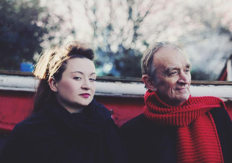 Eliza and Martin Carthy sitting in a boat outdoors. Eliza is looking at the camera and Martin is wearing a red scarf.