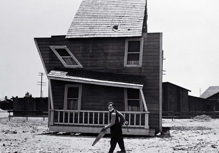 Buster Keaton stands with an saw outside a ramshackle, wonky wooden house