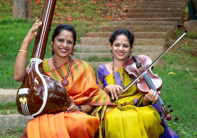 The Akkarai Sisters with their instruments, sitting on some steps in a garden