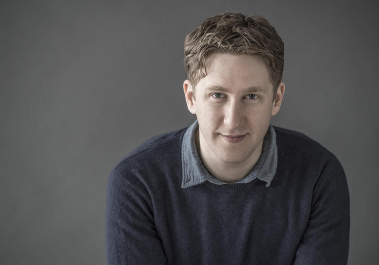 Conductor Joshua Weilerstein smiling at camera against a grey background