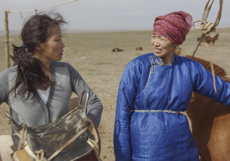 Two sisters in the desert look at one another, standing next to livestock