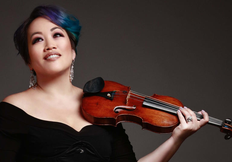 LSO First Violin Maxine Kwok smiles, holding a violin against a dark background