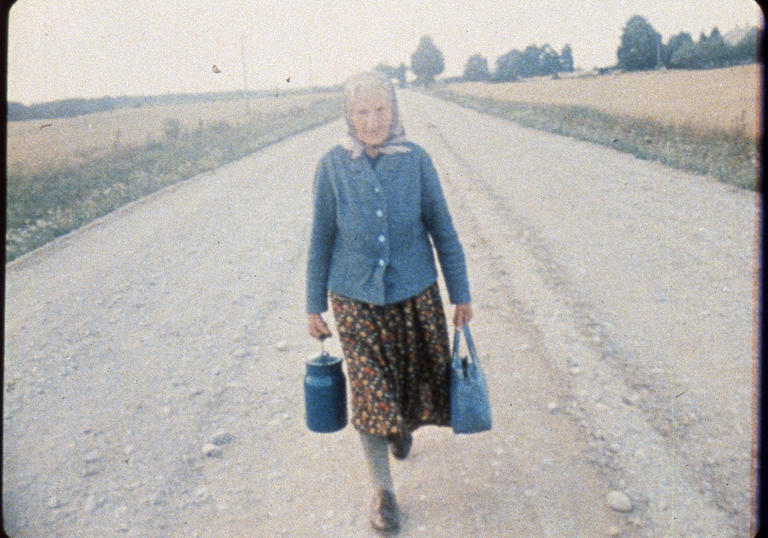 An elderly woman walks along an empty rural road in Jonas Mekas' Reminiscenes of a Journey to Lithuania