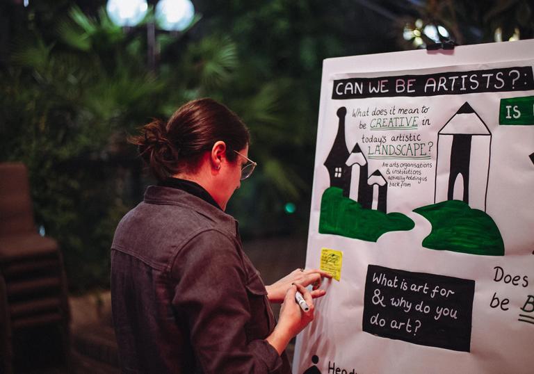 A person has their back to the camera and is writing on a large piece of paper which is hanging on a wall