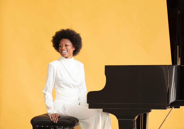 Isata Kanneh-Mason sitting at a grand piano smiling against a pale orange backdrop