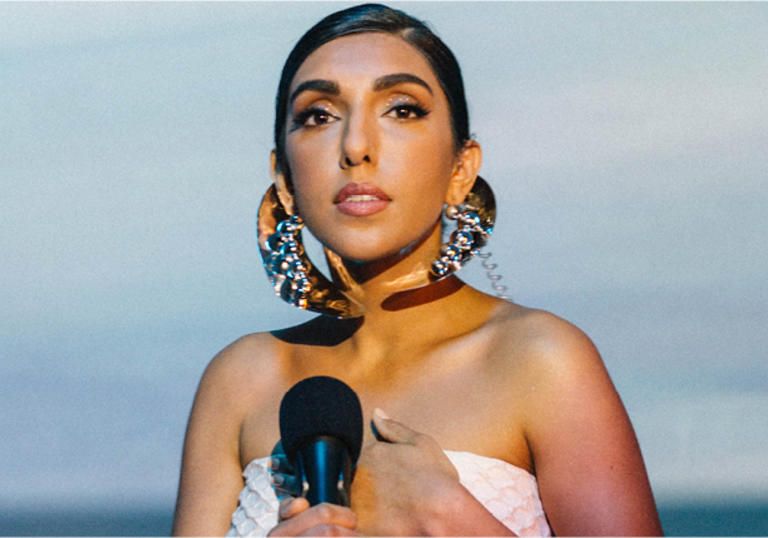 Rupi Kaur with gold hair decorations and jewellery, holding a microphone