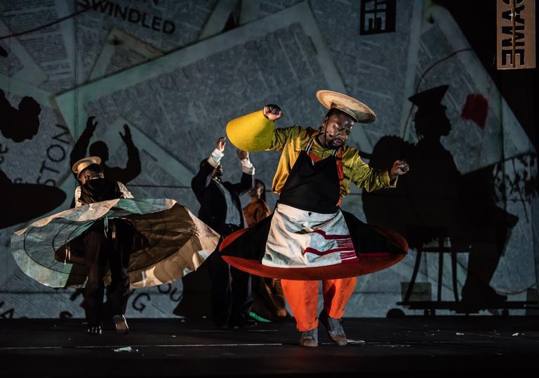 A person in a colourful and artistic costume dances, while other performers dance behind him. A projection of pages from a book lights up the back wall.