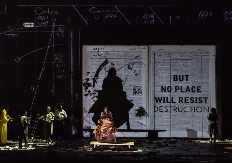 A performer in an orange dress dances in front of a large projection of a book. Other performers stand to the left-hand side and sing. 