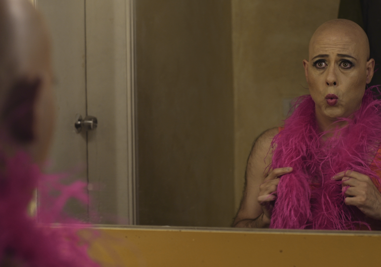 A person looks in the mirror with a pink feather boa in Miguel's War