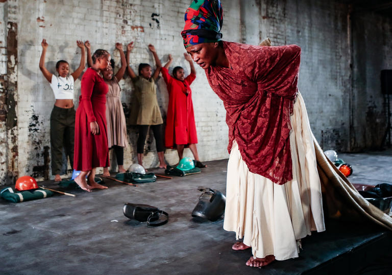 A performer in a cream dress and red shawl moves across the stage, behind her a group of 5 performers stand with their arms raised against the wall.