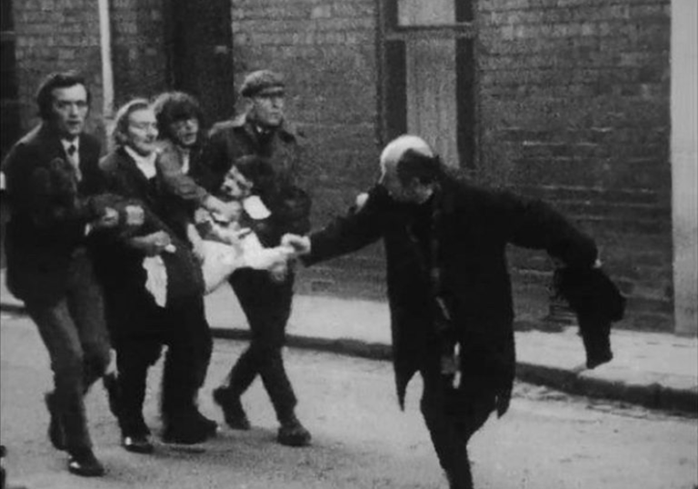 A group of men carry another injured man along the street in Ireland Behind the Wire