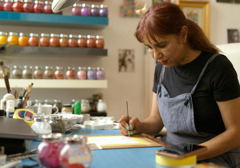 Artist Soheila is standing at her workstation in a blue apron and a paintbrush in her hand. On the wall behind are shelves with paint bottles.