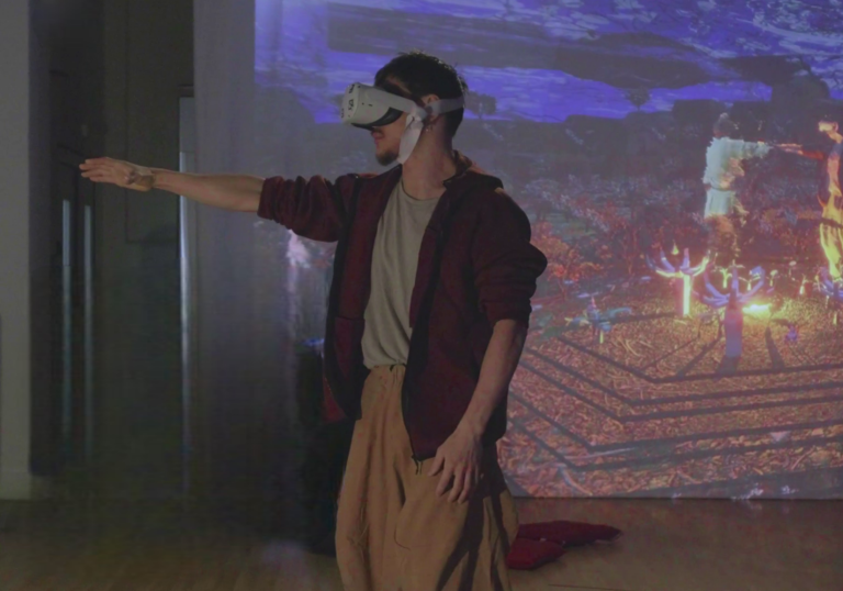 A person wearing a VR headset stands in front of a projector screen with their arm reaching towards something. On the projector screen the person appears in a 3D virtual world, reaching out to another person.