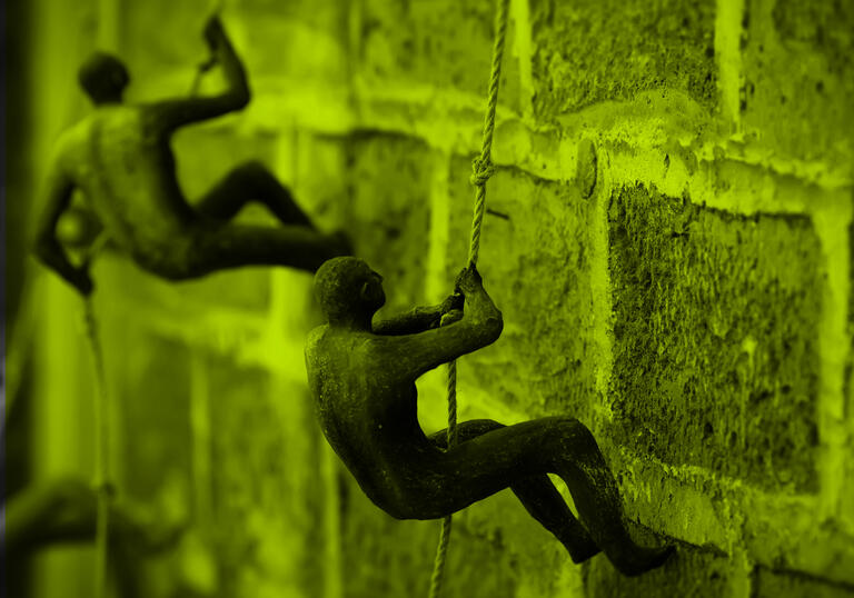 Two clay figurines climbing up brick wall using pieces of string