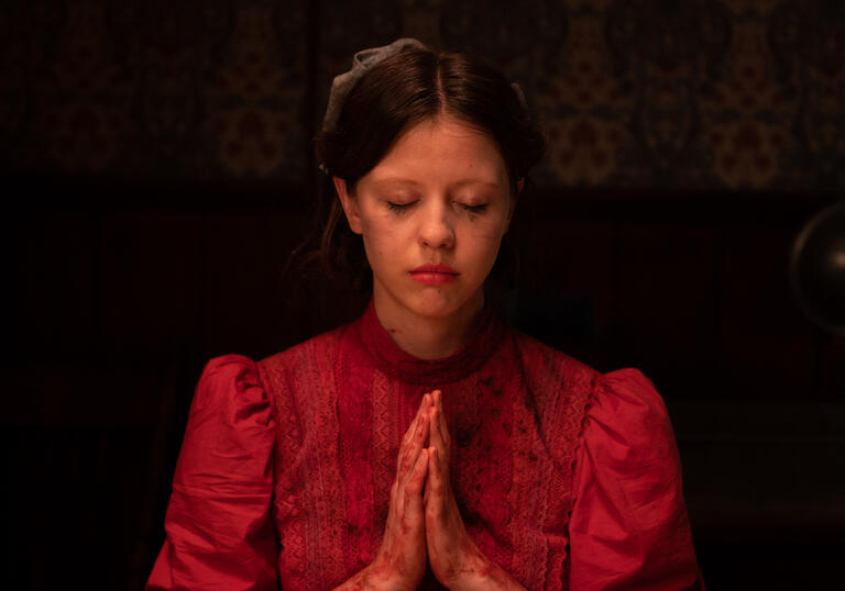 A young woman in a WWI era red dress prays, with running makeup as though she's been crying. A still from Pearl.