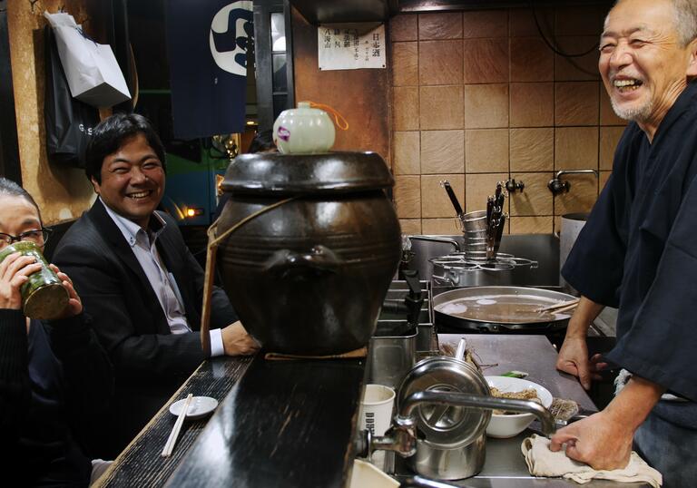 A smiling man stands in an open kitchen in a restaurant in front of customers in a still from Come Back Any Time