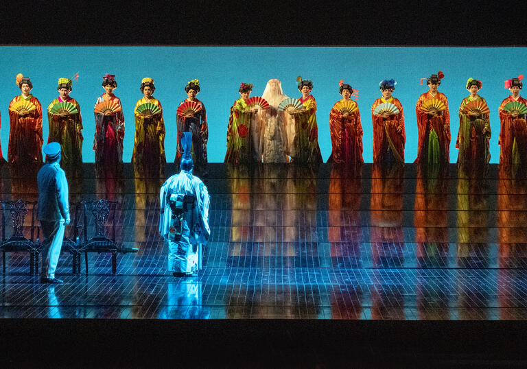 A row of people stand on a blue stage in traditional formal wear. 