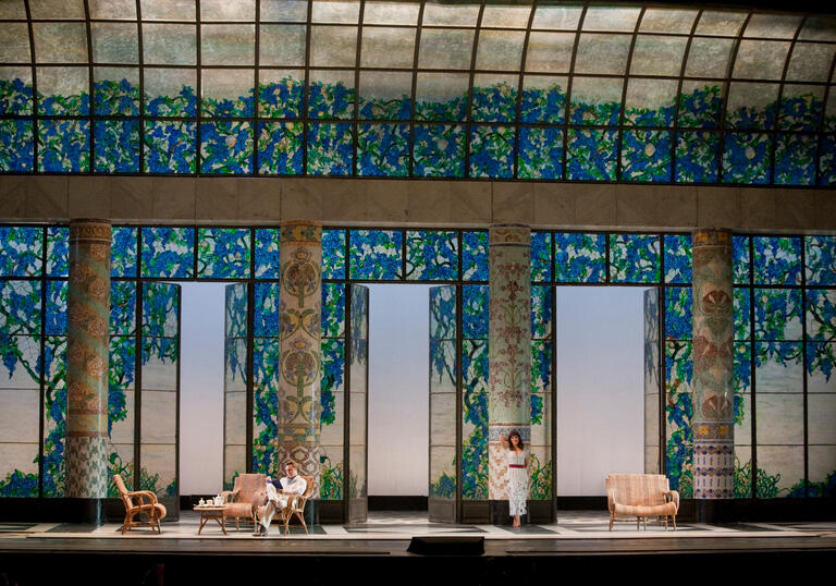 A man reads whilst a woman stands against a column on an ornate blue set on stage