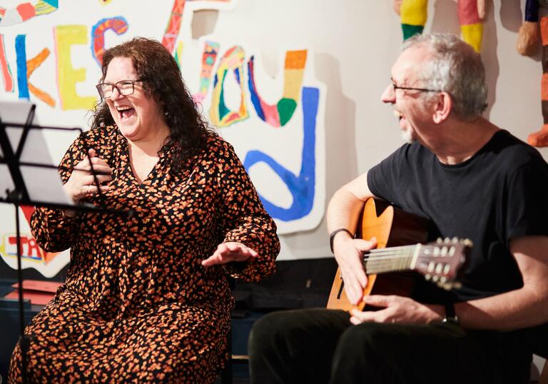 Two individuals, one is playing guitar and another looks on smiling