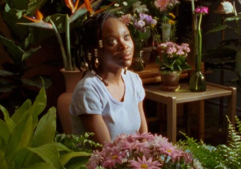 A woman looks to the side whilst surrounded by plants in a dream-like room