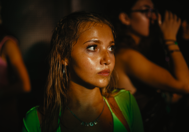 A young woman in a sweaty club looks forward, dazed, whilst bodies dance around her in the dark.