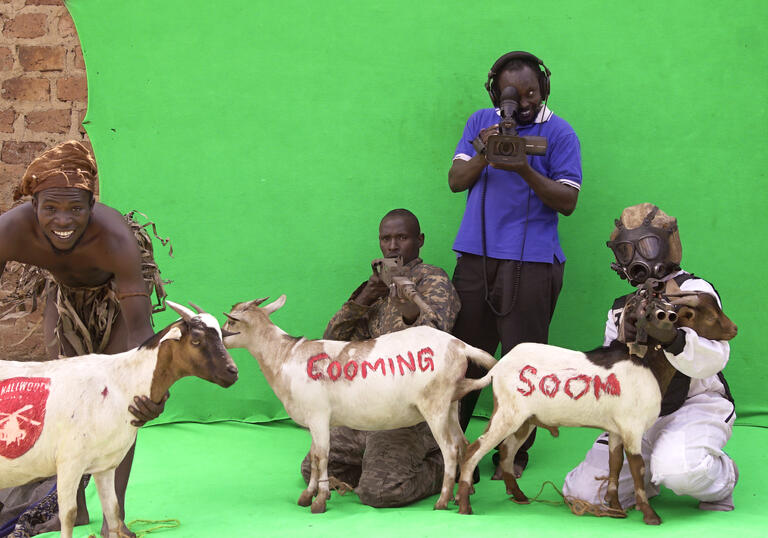 Two men sit on a green screen holding two goats, which have 'coming soon' written on their skin. A cameraman stands behind them. 