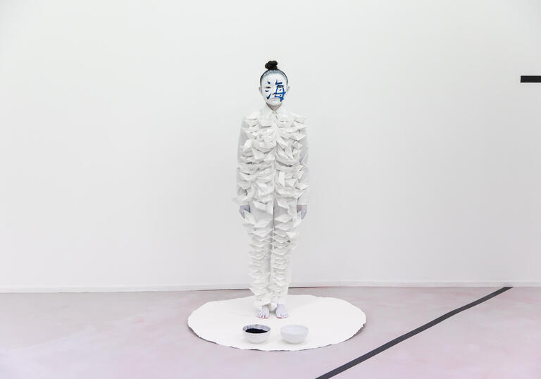 Performance artist Echo Morgan standing upright on a white fabric circle with two bowls of paint in front of her. She is wearing a paper bodysuit and her face is painted white and blue.