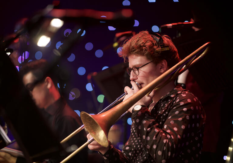 Musician playing trombone with headphones on and multicolour spotted pattern in background