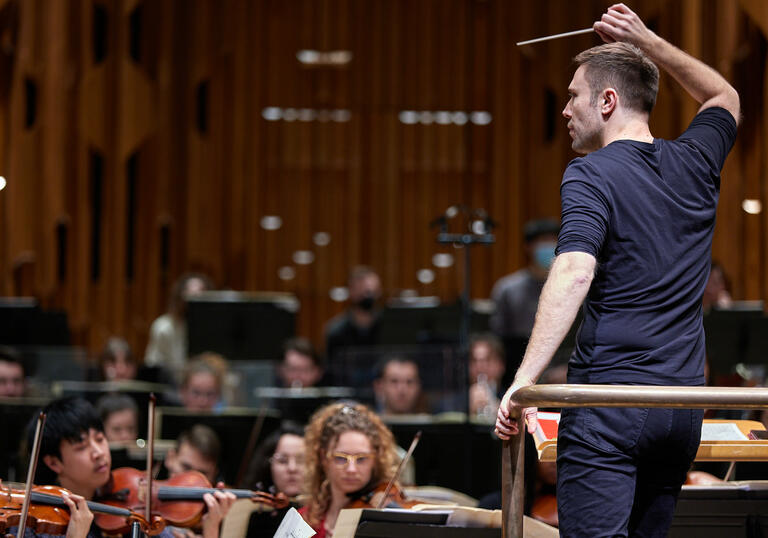 Conductor holding onto podium with baton in the air leading the violin section