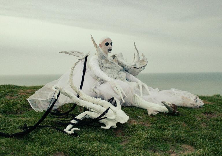 A performance artists lies on the grass of a clifftop, dressed in costume. 