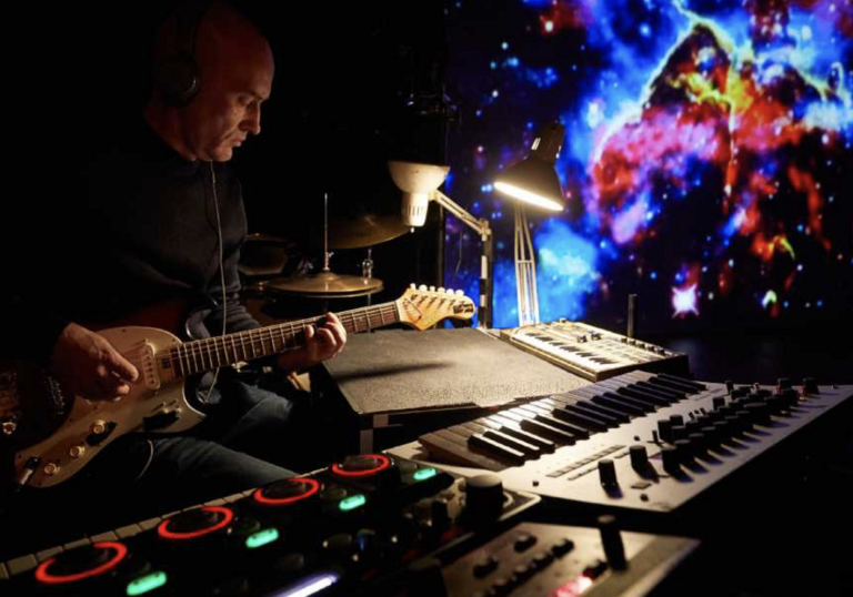 A person sits at a sound desk playing the guitar. Behind them, a projection of colours is on a screen.