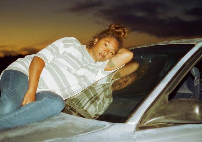 A young pregnant woman lies on the bonnet of a car in the dark, looking calmy forward. She wears blue jeans and a large striped teeshirt.