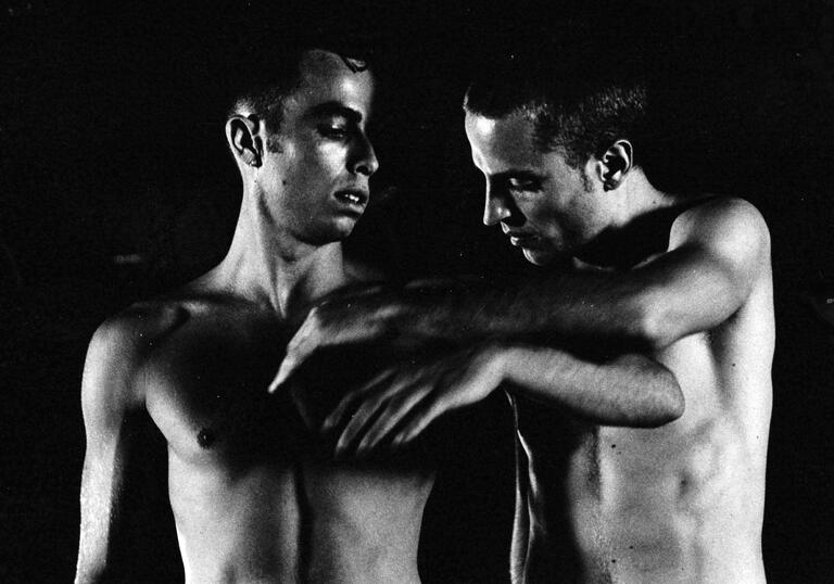 Two men stand topless in a dark room. 